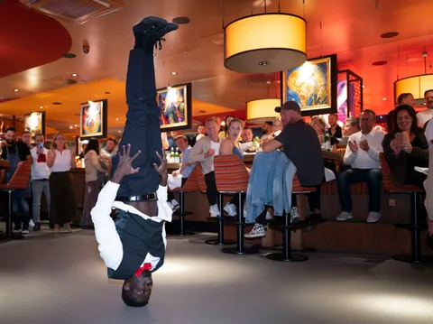 A show waiter performing a headstand in front of amazed and applauding restaurant guests.