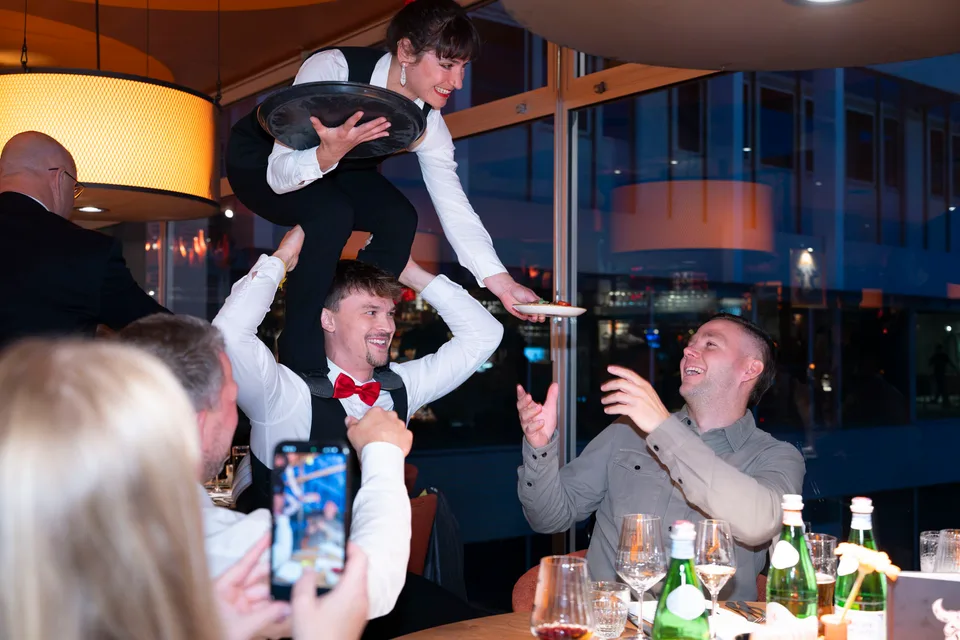 Acrobatics in waiter attire right at the table: a guest laughs while another records the moment on a phone.