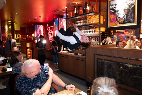 A show waiter performs a somersault beside restaurant tables while guests watch and film.