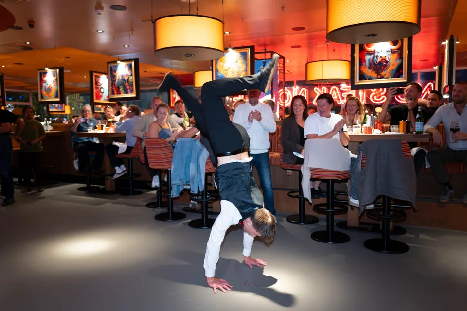 A show waiter performs a handstand in front of laughing and applauding guests.