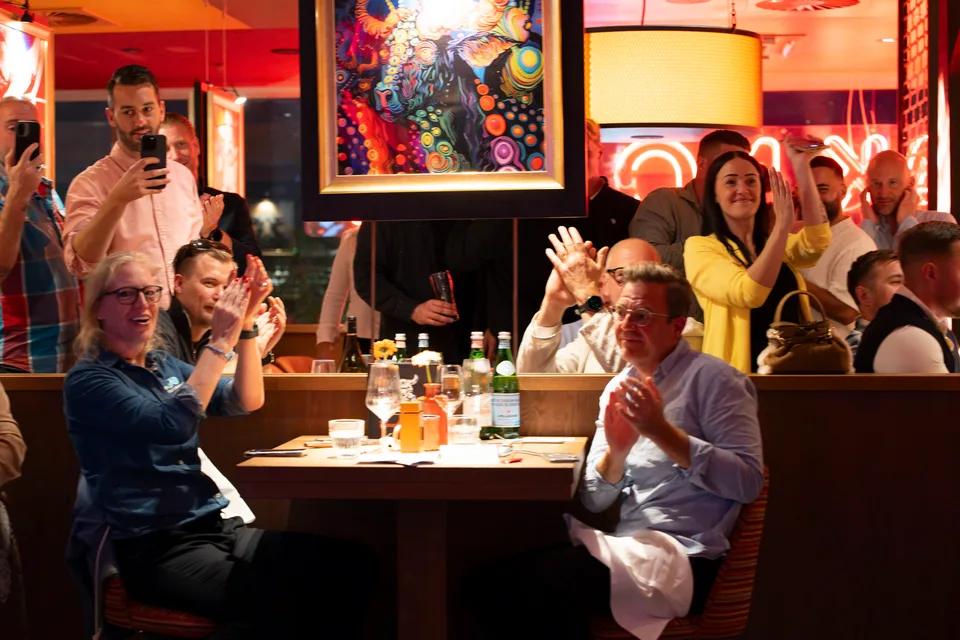 Guests applaud at restaurant tables and capture the show moment on their phones.