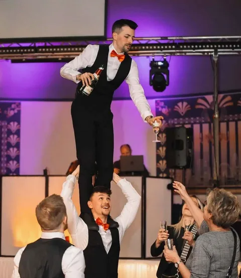A show waiter serves champagne while standing on his colleague’s shoulders.