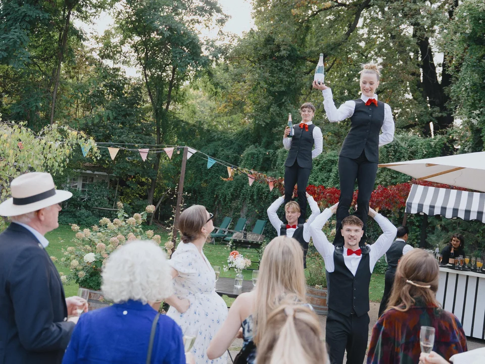 Two show waitresses stand on their colleagues’ shoulders among wedding guests in a garden.