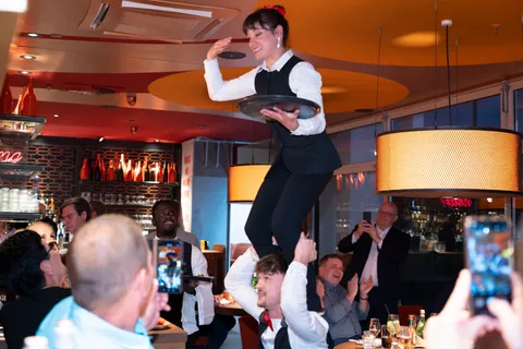 A show waitress holds a tray while standing on her colleague’s shoulders, surrounded by guests.