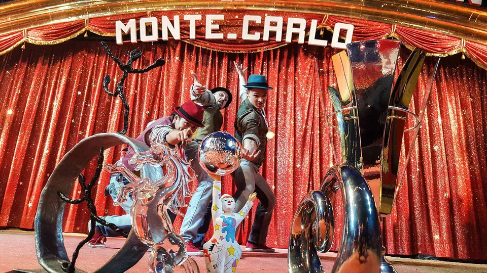 Three performers from The Funky Monkeys pose among trophies in front of a red curtain with a Monte-Carlo sign.